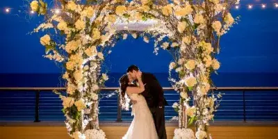 A wedding couple kissing under a large floral arch decorated with white roses and branches, set against a dark blue evening sky.