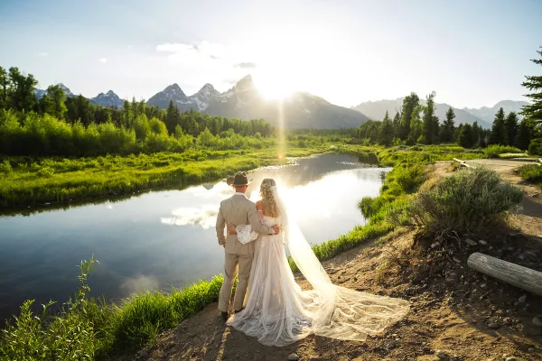 A bride and groom standing together in a scenic landscape with a river and mountains in the background.