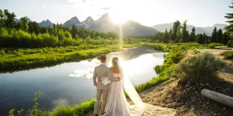 A bride and groom standing together in a scenic landscape with a river and mountains in the background.