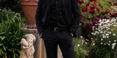 A man in a black suit standing in a garden with flowers and a statue in the background.