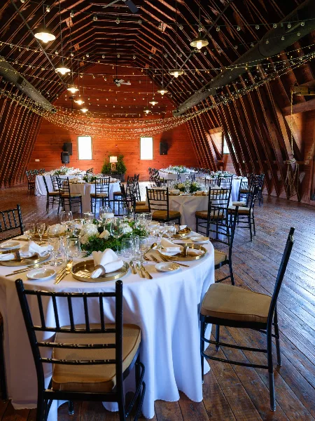 Interior of a timber hall with a high vaulted ceiling, string lights, and round tables set for a wedding reception.