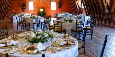 Interior of a timber hall with a high vaulted ceiling, string lights, and round tables set for a wedding reception.