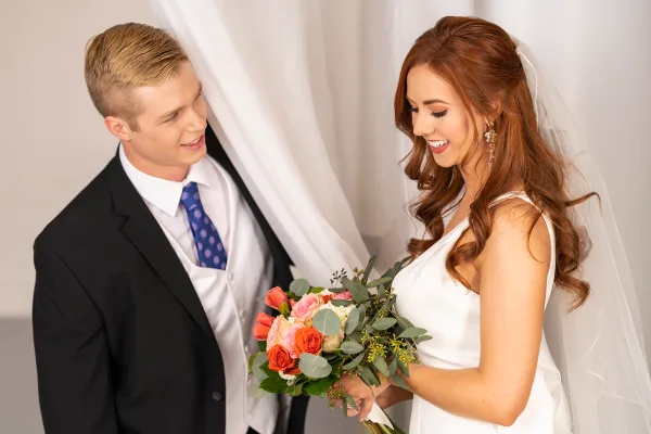 A bride and groom smiling at each other, with the bride holding a bouquet of pink and orange roses.