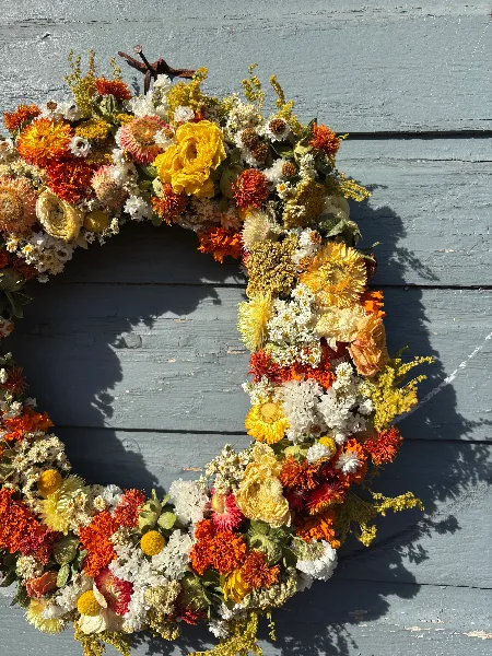 A large, lush floral wreath with orange, yellow, and white flowers hanging on a weathered blue wooden door.