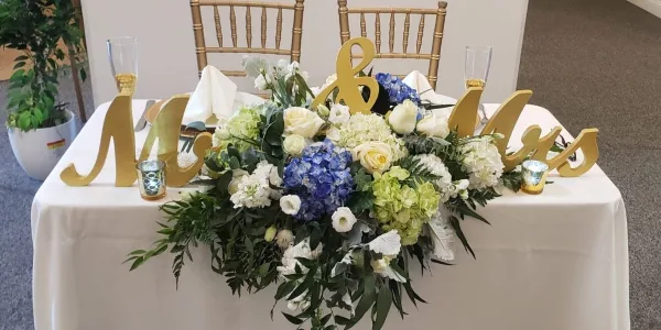 A wedding table centerpiece featuring white and blue flowers with greenery, accompanied by gold 'Mr & Mrs' letters.
