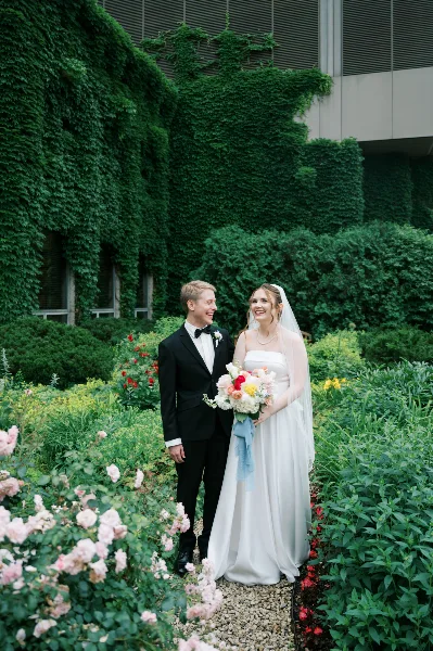 A bride and groom standing together in a garden setting, with the bride holding a bouquet.