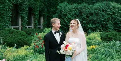 A bride and groom standing together in a garden setting, with the bride holding a bouquet.