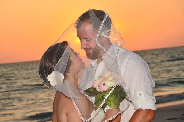 A bride and groom embracing on a beach at sunset, with the bride holding a bouquet of pink and white flowers.