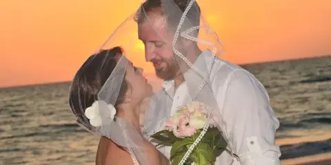 A bride and groom embracing on a beach at sunset, with the bride holding a bouquet of pink and white flowers.