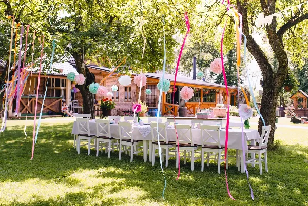 An outdoor event setup with white tables and chairs decorated with colorful streamers and paper pom-poms.