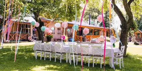 An outdoor event setup with white tables and chairs decorated with colorful streamers and paper pom-poms.