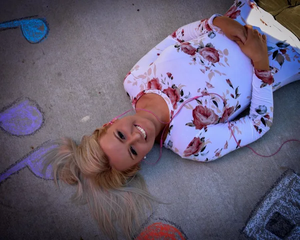 A smiling blonde woman in a floral top lying on a concrete surface with colorful chalk drawings.