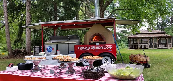 The Pizza Wagon mobile catering unit with a wood-fired oven and a table of prepared pizzas at an outdoor event.