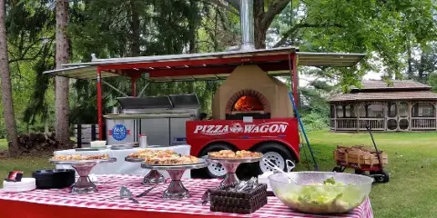 The Pizza Wagon mobile catering unit with a wood-fired oven and a table of prepared pizzas at an outdoor event.