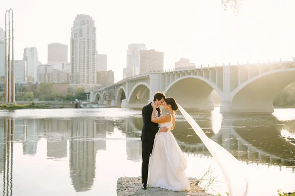 A bride and groom embracing by a river with a city skyline and bridge in the background.