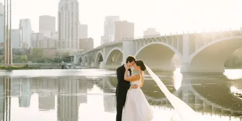 A bride and groom embracing by a river with a city skyline and bridge in the background.