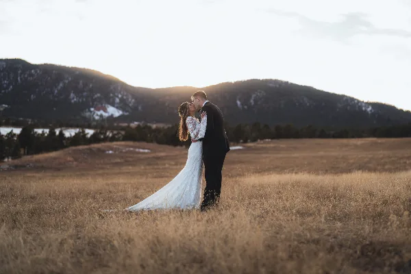 A bride and groom kissing in a grassy field with mountains in the background.