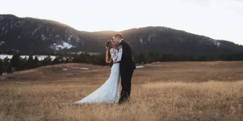 A bride and groom kissing in a grassy field with mountains in the background.