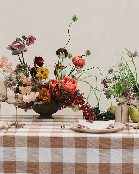 A lush mixed-flower centerpiece with red, orange, and pink blooms arranged in a dark bowl on a checkered tablecloth with glassware and fruit.