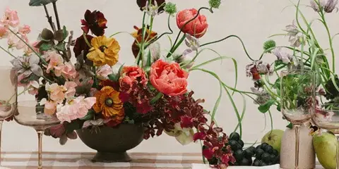 A lush mixed-flower centerpiece with red, orange, and pink blooms arranged in a dark bowl on a checkered tablecloth with glassware and fruit.