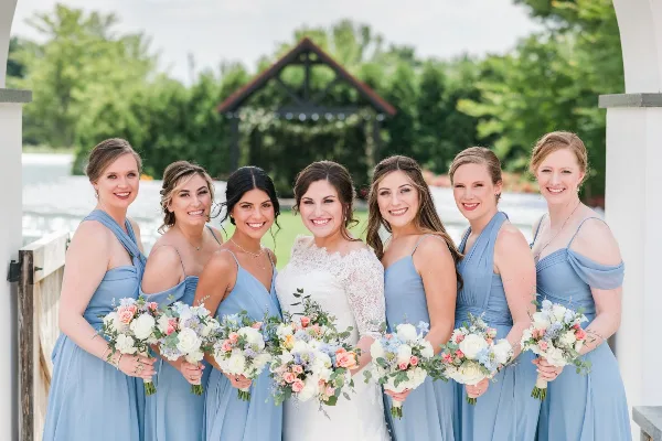 A bride in a white lace gown and six bridesmaids in blue dresses posing with bouquets, showcasing professional hair and makeup.
