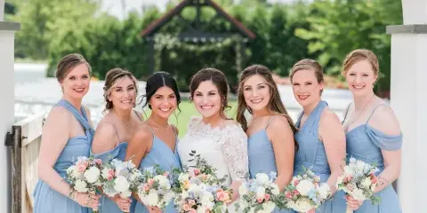 A bride in a white lace gown and six bridesmaids in blue dresses posing with bouquets, showcasing professional hair and makeup.