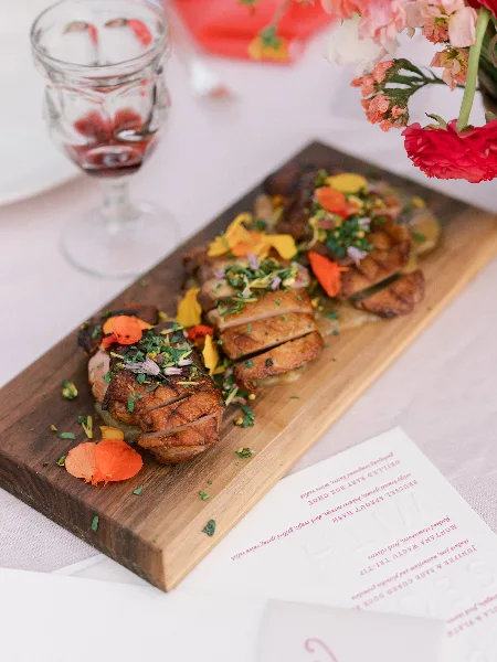 A plated dish of sliced roasted meat with garnishes and edible flowers on a wooden serving board, accompanied by a glass of red wine and a floral arrangement.