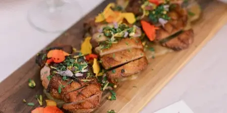 A plated dish of sliced roasted meat with garnishes and edible flowers on a wooden serving board, accompanied by a glass of red wine and a floral arrangement.