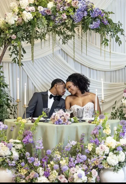 Newlywed couple seated at a reception table beneath a large floral installation of purple and white blooms.
