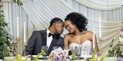Newlywed couple seated at a reception table beneath a large floral installation of purple and white blooms.