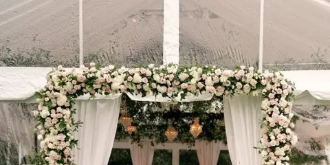 A wedding reception setup inside a white tent featuring a floral arch and round tables.