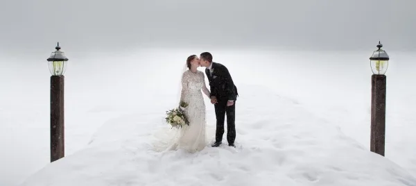 A bride and groom kissing in a snowy landscape, flanked by two tall wooden posts with lanterns.