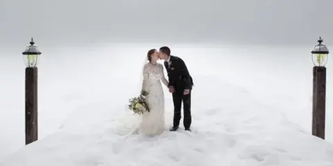 A bride and groom kissing in a snowy landscape, flanked by two tall wooden posts with lanterns.