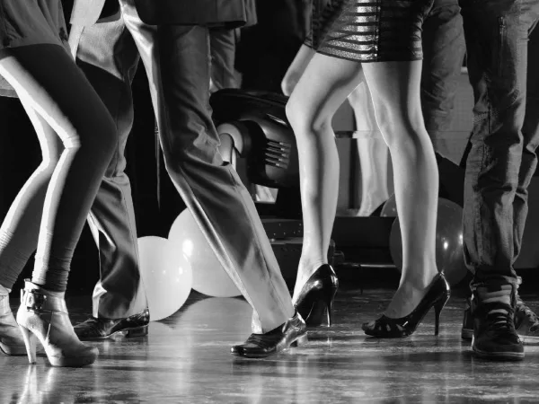 A black and white photograph showing the legs and feet of several people dancing at a party, with balloons and event lighting visible in the background.