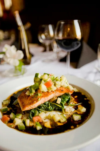 A plated salmon dish with a vegetable garnish served on a white tablecloth with wine glasses in the background.
