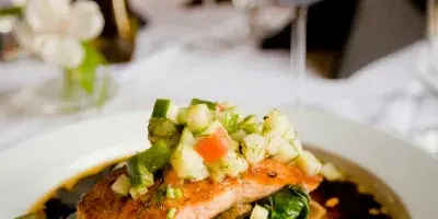 A plated salmon dish with a vegetable garnish served on a white tablecloth with wine glasses in the background.