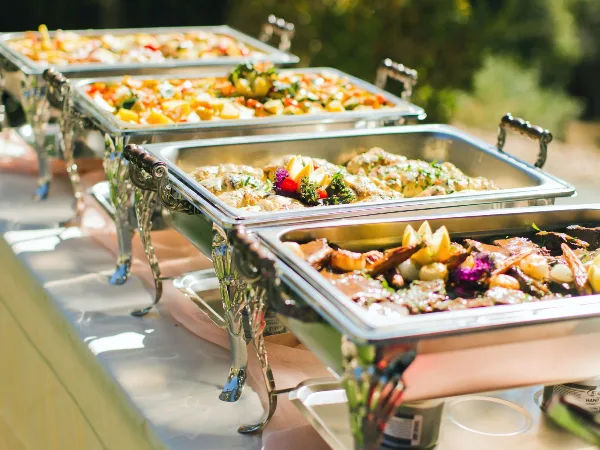 A row of silver chafing dishes filled with various prepared food items, including salads and roasted meats, set up for a catering event.
