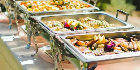 A row of silver chafing dishes filled with various prepared food items, including salads and roasted meats, set up for a catering event.