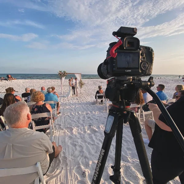 A professional video camera on a tripod recording a beach wedding ceremony with a bride and groom at an altar.