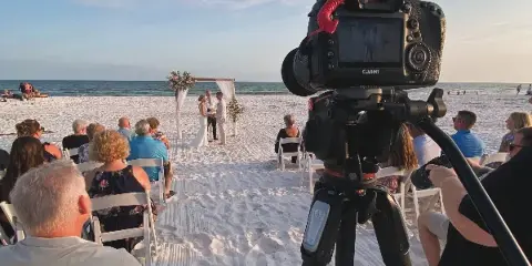A professional video camera on a tripod recording a beach wedding ceremony with a bride and groom at an altar.
