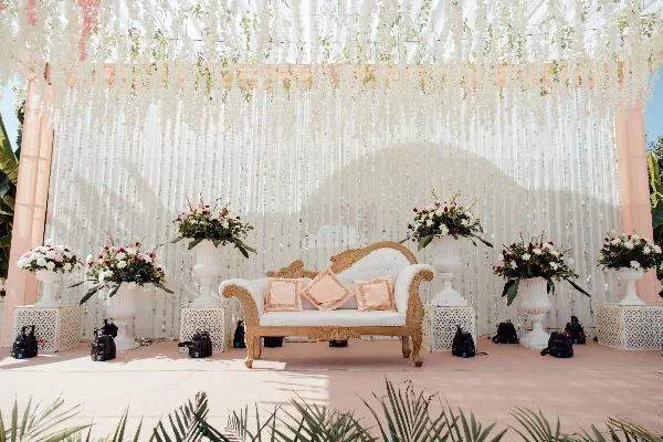 A wedding stage setup featuring a white and gold sofa, floral arrangements in white urns, and a beaded backdrop.