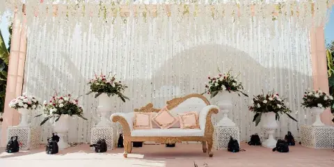 A wedding stage setup featuring a white and gold sofa, floral arrangements in white urns, and a beaded backdrop.