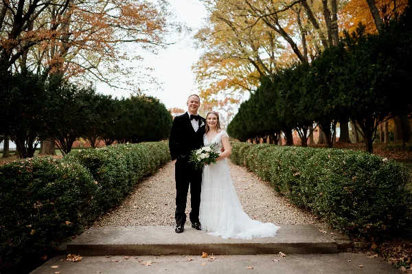 A bride and groom standing together on a garden path lined with hedges and autumn trees.