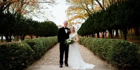 A bride and groom standing together on a garden path lined with hedges and autumn trees.