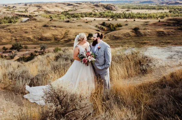 Bride and groom standing together in tall grass on a hillside overlooking rolling North Dakota prairie hills.