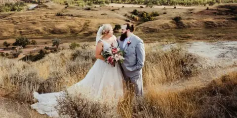 Bride and groom standing together in tall grass on a hillside overlooking rolling North Dakota prairie hills.