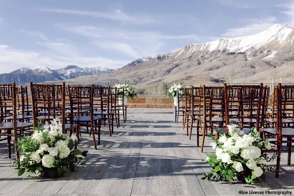 Outdoor wedding ceremony seating with white floral arrangements on a wooden deck, set against a backdrop of snow-capped mountains.