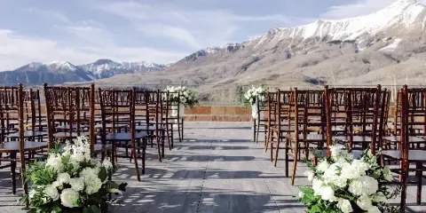 Outdoor wedding ceremony seating with white floral arrangements on a wooden deck, set against a backdrop of snow-capped mountains.