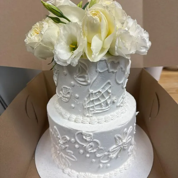 A two-tier white wedding cake with white buttercream icing, decorative piping, and a bouquet of white flowers on top.