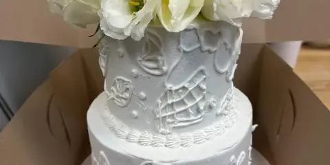 A two-tier white wedding cake with white buttercream icing, decorative piping, and a bouquet of white flowers on top.